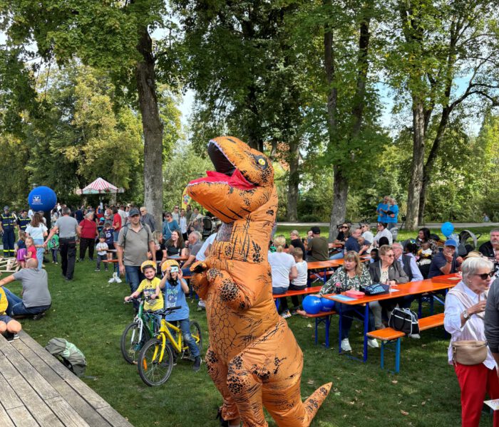 Bild des Mannes im Dinosaurier-Kostüm im Hintergrund Besucher*innen auf Bierbänken